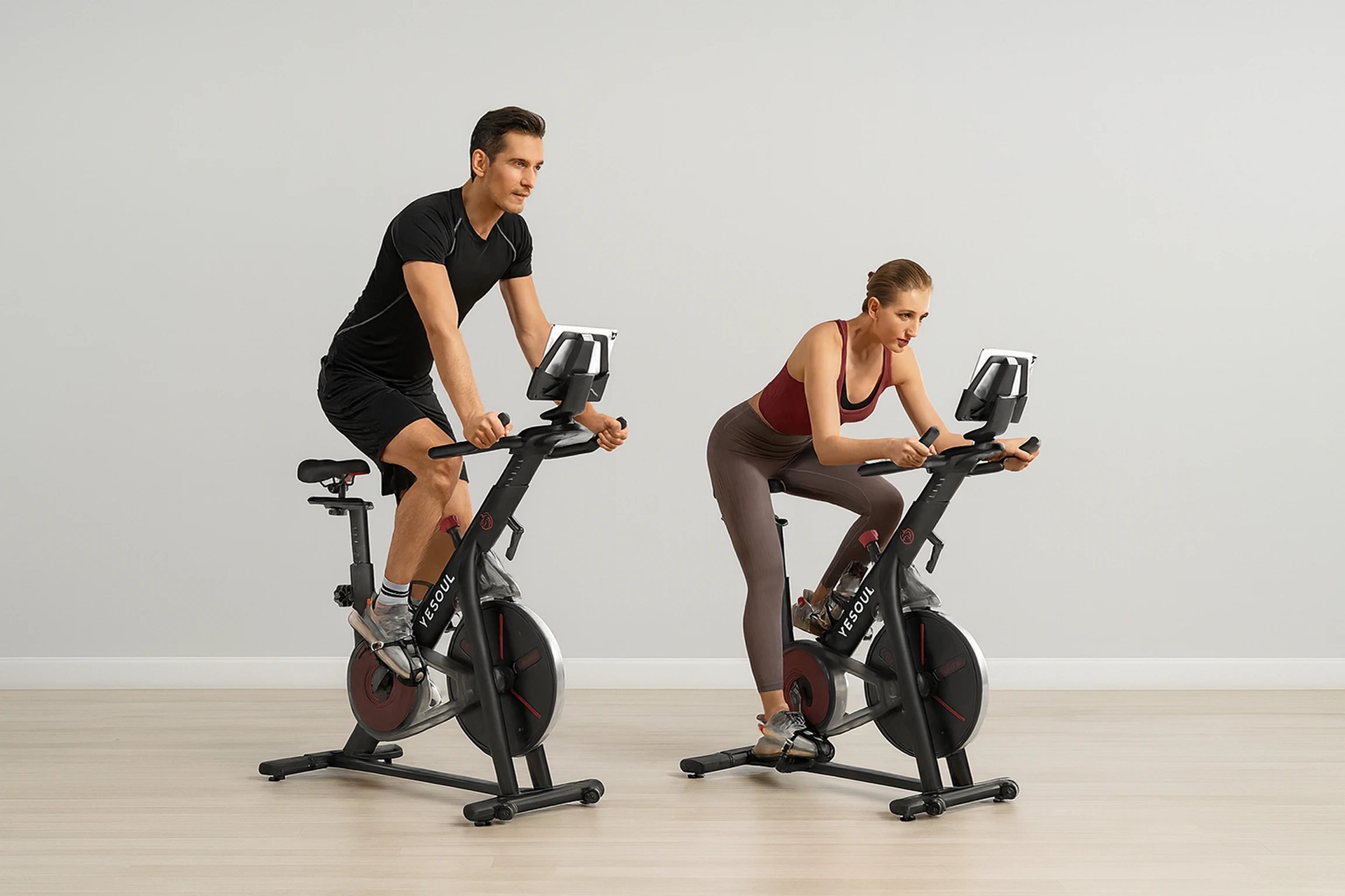 Due persone si allenano su bike indoor Yesoul con un tablet appoggiato sul porta-telefono per un allenamento smart, in un ambiente interno minimal e luminoso